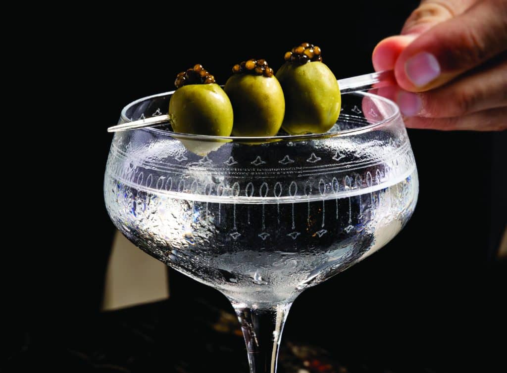 A person grips a knife in one hand and a glass filled with olives in the other, showcasing a culinary preparation moment.