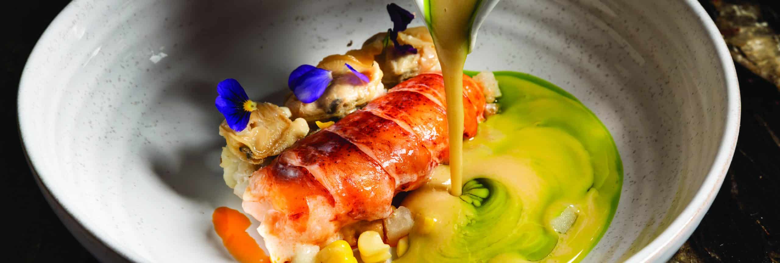 A lobster on a white plate is being drizzled with a rich sauce, enhancing its vibrant color and presentation.